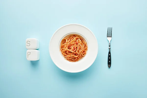 Flat lay with delicious spaghetti with tomato sauce near fork, salt and pepper shakers on blue background — Stock Photo