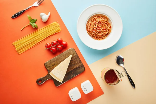Plat étendre avec de délicieux spaghettis à la sauce tomate dans une assiette près de couverts et ingrédients sur fond bleu, rouge et jaune — Photo de stock