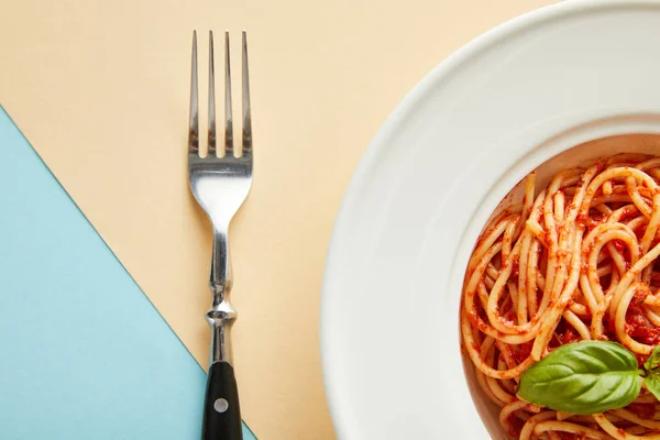 Вид сверху на вкусные спагетти с томатным соусом в тарелке рядом с вилкой на синем и желтом фоне — стоковое фото