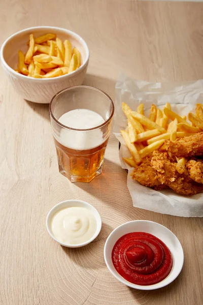 Vaso de cerveza, nuggets de pollo con papas fritas, ketchup y mayonesa sobre mesa de madera - foto de stock