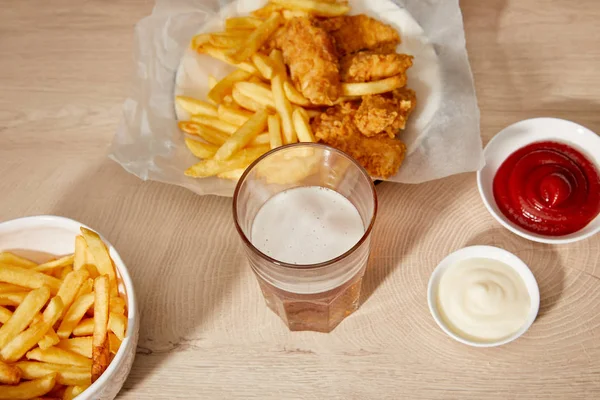 Vaso de cerveza, nuggets de pollo con papas fritas, ketchup y mayonesa sobre mesa de madera - foto de stock