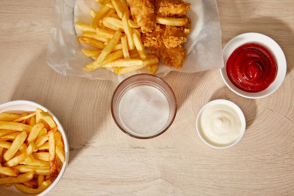 Vista superior de vidro de cerveja, nuggets de frango com batatas fritas, ketchup e maionese na mesa de madeira — Fotografia de Stock