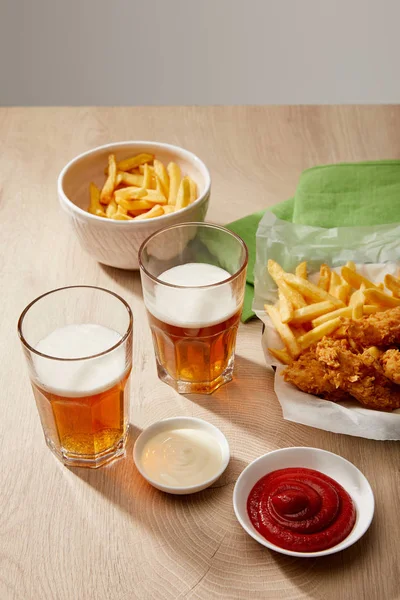 Glasses of beer, chicken nuggets with french fries, ketchup and mayonnaise on wooden table on grey background — Stock Photo