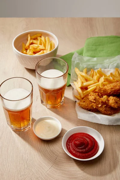 Glasses of beer, chicken nuggets with french fries, ketchup and mayonnaise on wooden table on grey background — Stock Photo