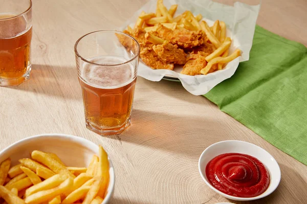 Vasos de cerveza, nuggets de pollo con papas fritas, ketchup sobre mesa de madera - foto de stock