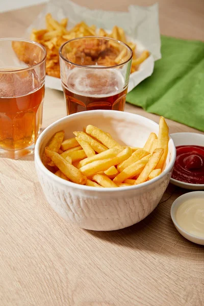 Bicchieri di birra, patatine fritte in ciotola, salse sul tavolo di legno — Foto stock