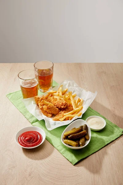 Vasos de cerveza, pepitas de pollo con papas fritas, salsas y pepinillos sobre mesa de madera sobre fondo gris - foto de stock
