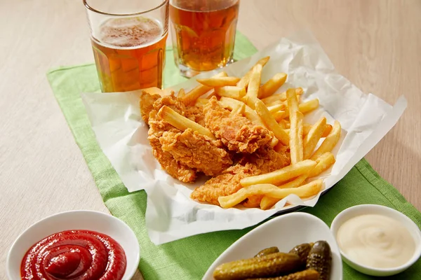 Verres de bière, pépites de poulet avec frites, sauces et cornichons sur table en bois — Photo de stock