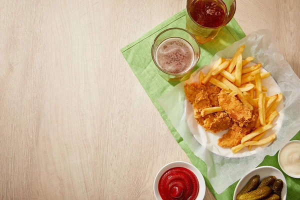 Vista superior de copos de cerveja, nuggets de frango com batatas fritas, molhos e pepinos em mesa de madeira com espaço de cópia — Fotografia de Stock