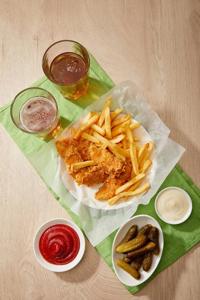 Verres de bière, pépites de poulet avec frites, sauces et cornichons sur table en bois — Photo de stock