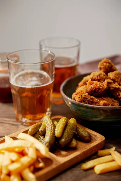 Селективный фокус вкусных куриных наггетсов, картошки фри и огурцов возле бокалов пива на деревянном столе, изолированном на сером — стоковое фото