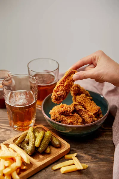 Vista recortada de la mujer comiendo deliciosas pepitas de pollo, papas fritas y pepinillos cerca de vasos de cerveza en una mesa de madera aislada en gris - foto de stock