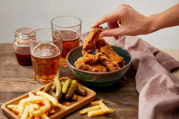 Vista recortada de la mujer comiendo deliciosas pepitas de pollo, papas fritas y pepinillos cerca de vasos de cerveza en una mesa de madera aislada en gris - foto de stock