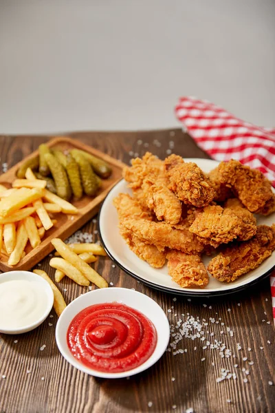 Delicious chicken nuggets, ketchup, mayonnaise, french fries and gherkins on wooden table with salt and rustic plaid napkin isolated on grey — Stock Photo