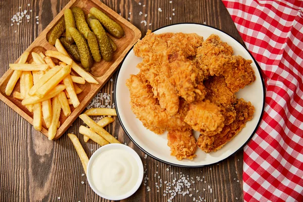 Vista superior de deliciosas pepitas de pollo, mayonesa, papas fritas y pepinillos en mesa de madera con sal y servilleta a cuadros rústica - foto de stock