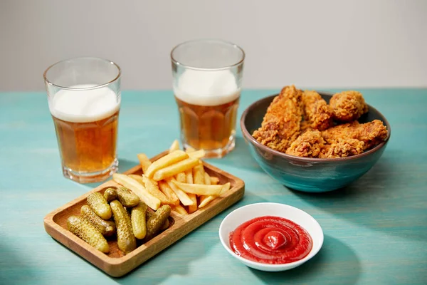Deliziose pepite di pollo, ketchup, patatine fritte e cetriolini vicino a bicchieri di birra su tavolo di legno turchese isolato su grigio — Foto stock