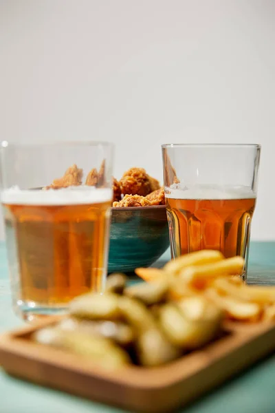 Enfoque selectivo de deliciosas pepitas de pollo, papas fritas y pepinillos cerca de vasos de cerveza sobre mesa de madera turquesa sobre fondo gris - foto de stock