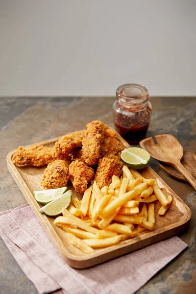 Planche de bois avec de délicieuses pépites de poulet, frites et citron vert sur la surface de pierre isolée sur gris — Photo de stock