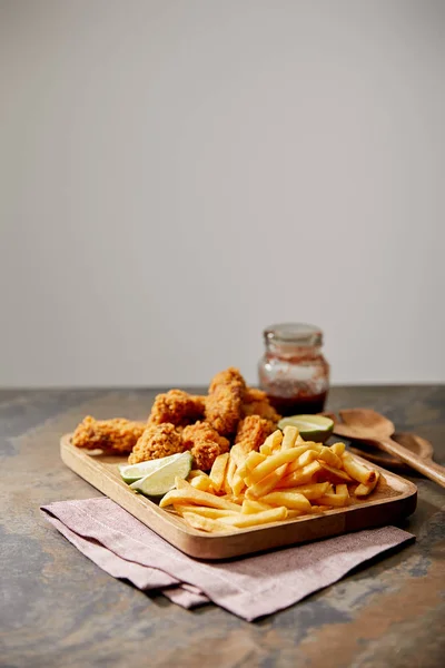 Planche de bois avec de délicieuses pépites de poulet, frites et citron vert sur la surface de pierre isolée sur gris — Photo de stock