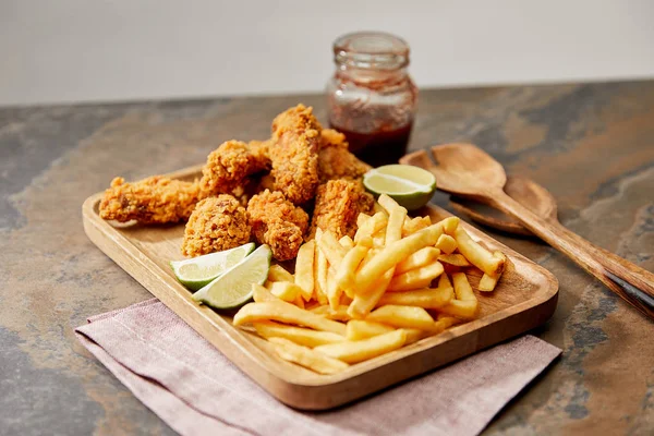 Bordo di legno con deliziose pepite di pollo, patatine fritte e lime su superficie in pietra isolata su grigio — Foto stock