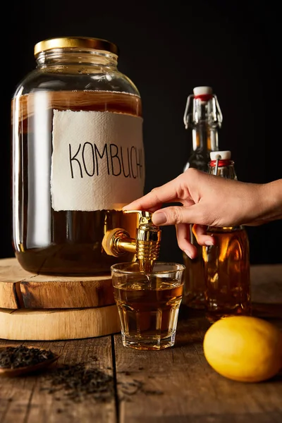Cropped view of woman pouring kombucha in glass from jar on wooden table isolated on black — Stock Photo