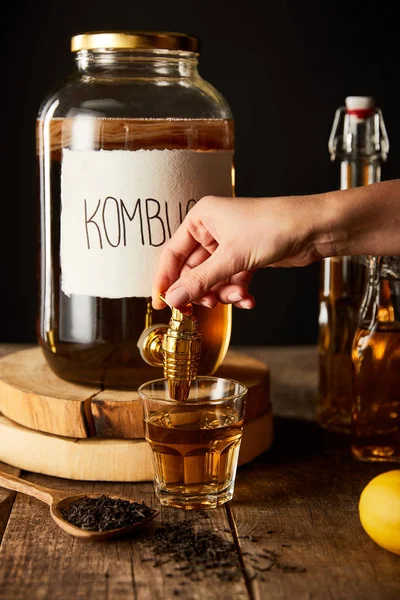 Cropped view of woman pouring kombucha in glass from jar on wooden table isolated on black — Stock Photo