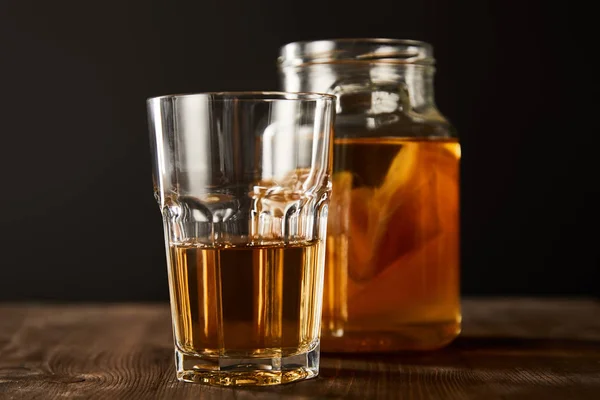 Selective focus of glass and jar with kombucha on wooden table isolated on black — Stock Photo