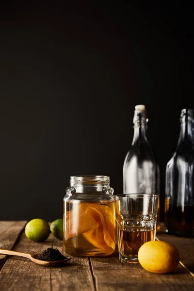 Glass jar with kombucha near lime, lemon, spice and bottles on wooden table isolated on black — Stock Photo