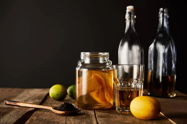 Bocal en verre avec kombucha près de citron vert, citron, épices et bouteilles sur table en bois isolé sur noir — Photo de stock