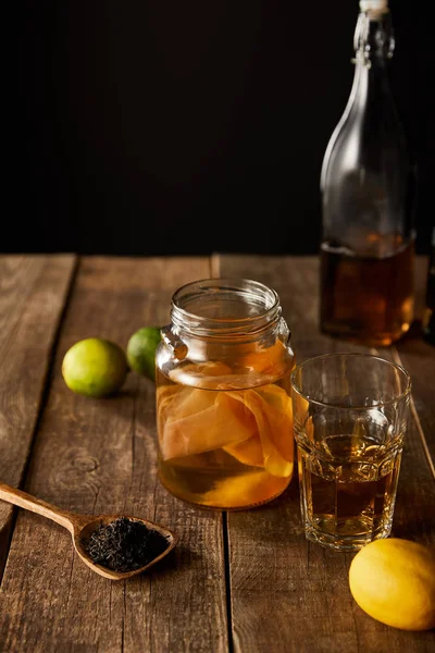 Selective focus of glass jar with kombucha near lime, lemon, spice and bottle on wooden table isolated on black — Stock Photo