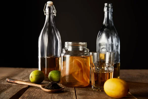 Vaso di vetro con kombucha vicino a lime, limone, spezie e bottiglie su tavolo di legno isolato su nero — Foto stock