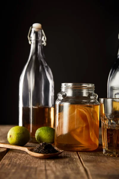 Glass jar with kombucha near lime, spice and bottle on wooden table isolated on black — Stock Photo