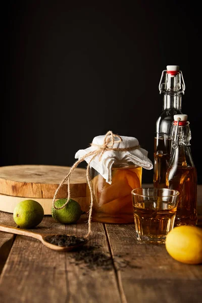 Glass jar with kombucha near lime, lemon, spice and bottles on wooden table isolated on black — Stock Photo