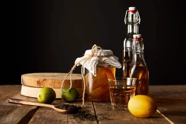 Vaso di vetro con kombucha vicino a lime, limone, spezie e bottiglie su tavolo di legno isolato su nero — Foto stock