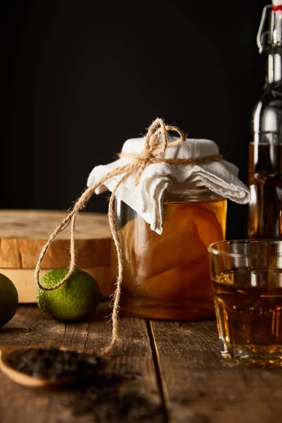 Selective focus of glass jar with kombucha near lime, spice and bottle on wooden table isolated on black — Stock Photo