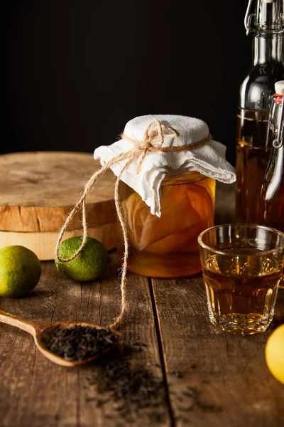 Vaso di vetro con kombucha vicino lime, limone, spezie e bottiglia su tavolo di legno isolato su nero — Foto stock