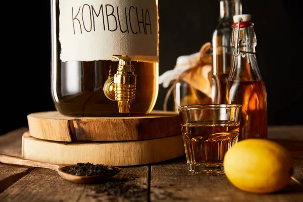 Vista de cerca del frasco de vidrio con kombucha cerca de limón, especias y botellas en mesa de madera aislada en negro - foto de stock