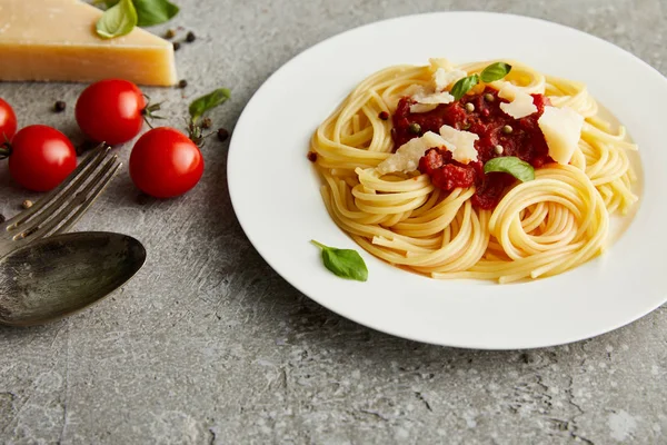 Gustosa pasta bolognese con sugo di pomodoro e parmigiano su piatto bianco vicino agli ingredienti e posate su fondo grigio — Foto stock