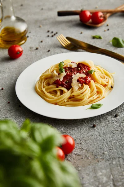 Foco seletivo de massas bolonhesas saborosas com molho de tomate e parmesão em prato branco perto de ingredientes e talheres em fundo cinza — Fotografia de Stock