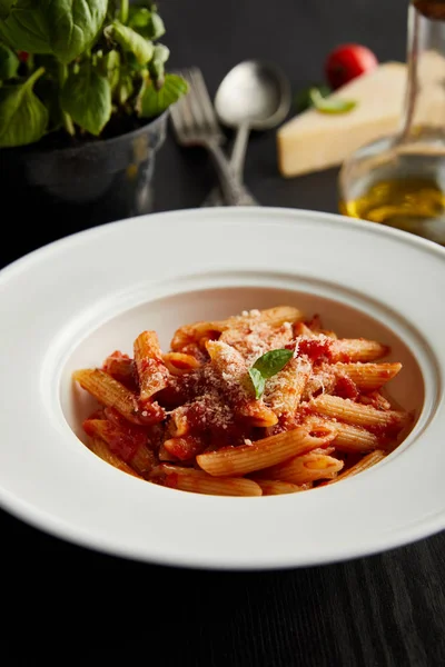 Messa a fuoco selettiva di gustosa pasta bolognese con sugo di pomodoro e parmigiano su piatto bianco vicino agli ingredienti e posate su fondo nero in legno — Foto stock