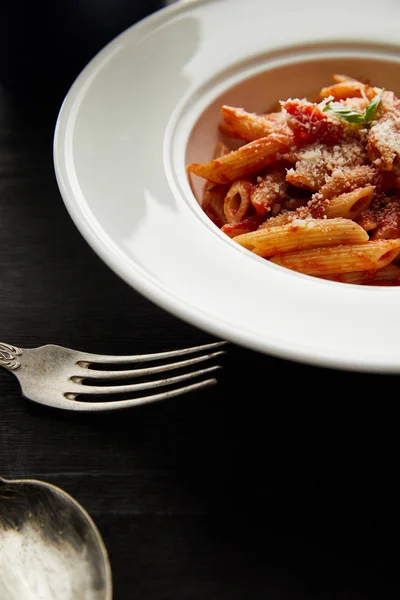 Tasty bolognese pasta with tomato sauce and Parmesan in white plate near cutlery on black wooden background — Stock Photo