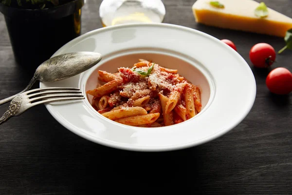 Foco seletivo de massa bolonhesa saborosa com molho de tomate e parmesão em placa branca perto de ingredientes e talheres em fundo de madeira preta — Fotografia de Stock