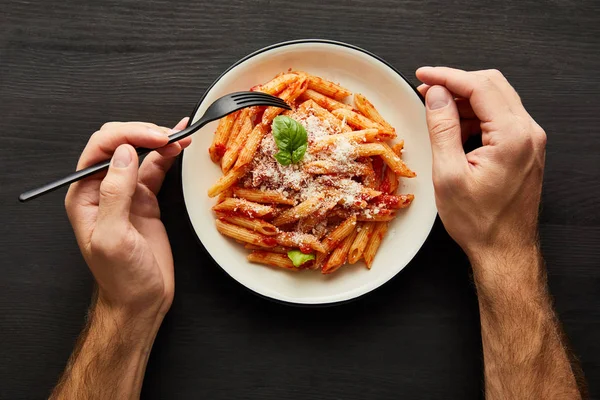 Cropped view of man eating tasty bolognese pasta with tomato sauce and Parmesan from white plate on black wooden background — Stock Photo