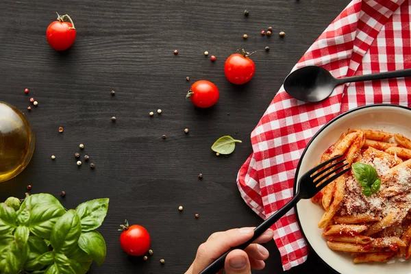 Vue recadrée de l'homme mangeant de savoureuses pâtes bolognaises à la sauce tomate et le parmesan de plaque blanche sur table en bois noir avec des ingrédients et serviette à carreaux — Photo de stock