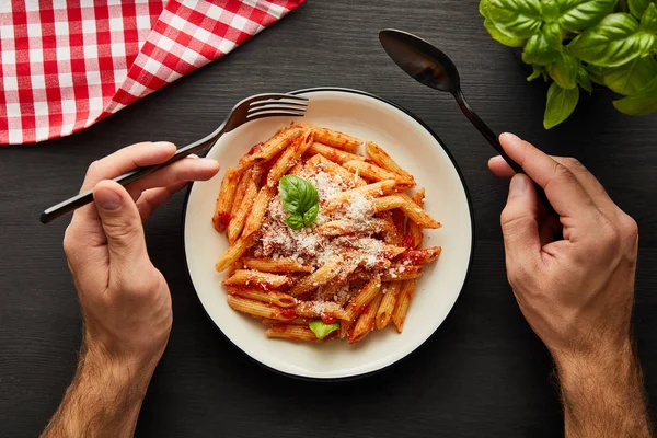 Abgeschnittene Ansicht des Mannes, der leckere Bolognese-Pasta mit Tomatensauce und Parmesan von weißem Teller auf schwarzem Holztisch mit Basilikum und Karo-Serviette isst — Stockfoto