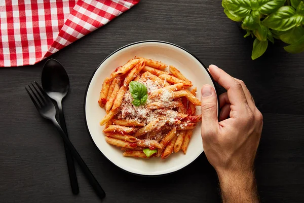 Cropped view of man holding plate with tasty bolognese pasta on black wooden table with basil, cutlery and check napkin — Stock Photo