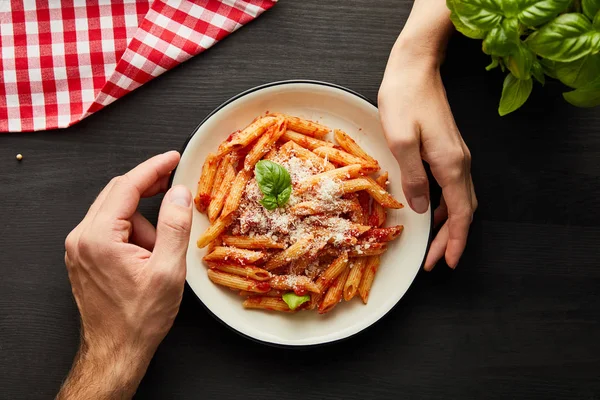 Vue recadrée du couple tenant assiette avec de savoureuses pâtes bolognaises sur table en bois noir avec basilic, couverts et serviette à carreaux — Photo de stock