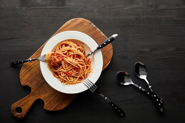 Vue du dessus de savoureuses pâtes bolognaises avec sauce tomate dans une assiette blanche sur planche à découper en bois près des couverts sur fond en bois noir — Photo de stock
