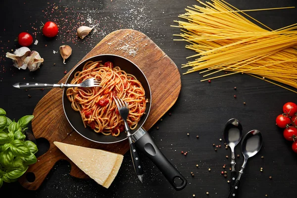 Top view of tasty bolognese pasta in frying pan near ingredients and cutlery on black wooden background — Stock Photo