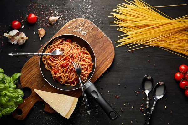 Vista superior de la deliciosa pasta boloñesa en sartén cerca de ingredientes y cubiertos sobre fondo de madera negro - foto de stock